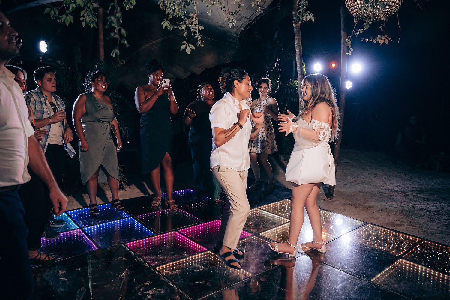 Wedding guests dance on an illuminated dance floor at night during an outdoor celebration.