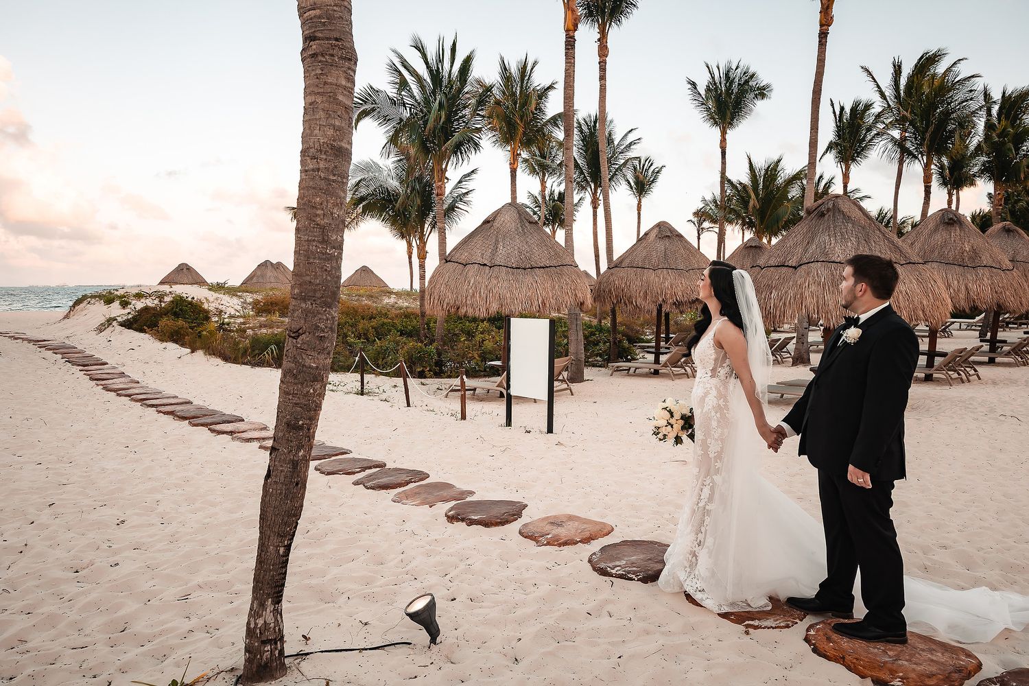 Romantic beach wedding ceremony with palm trees and thatched umbrellas at sunset in a tropical destination.