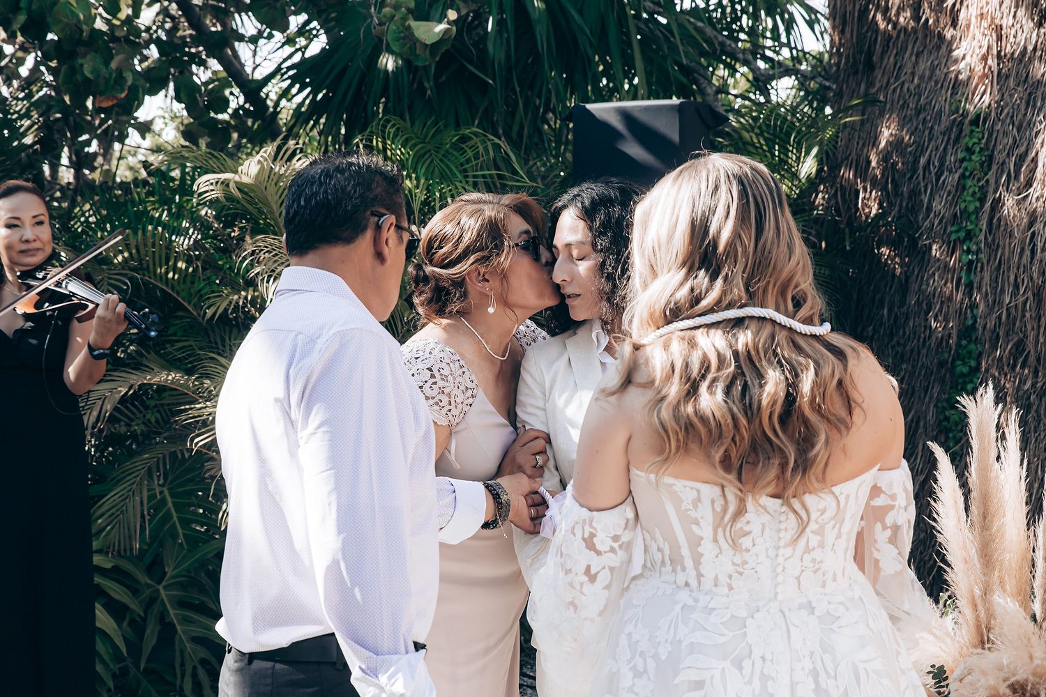 A wedding party gathers outdoors in a garden setting, with people dressed in white and beige attire.