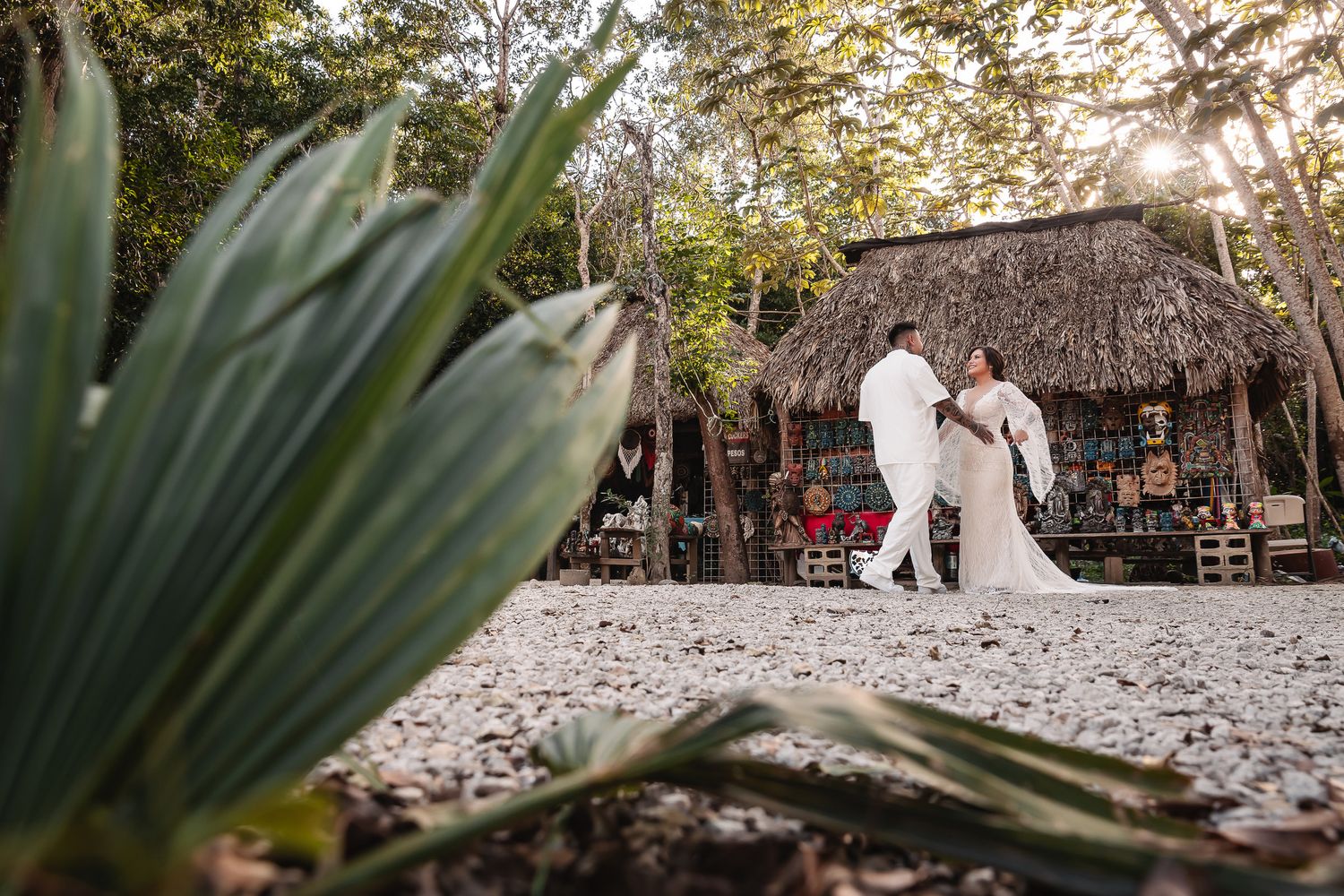 A romantic beach wedding ceremony takes place at sunset near a thatched-roof palapa with palm fronds in the foreground.