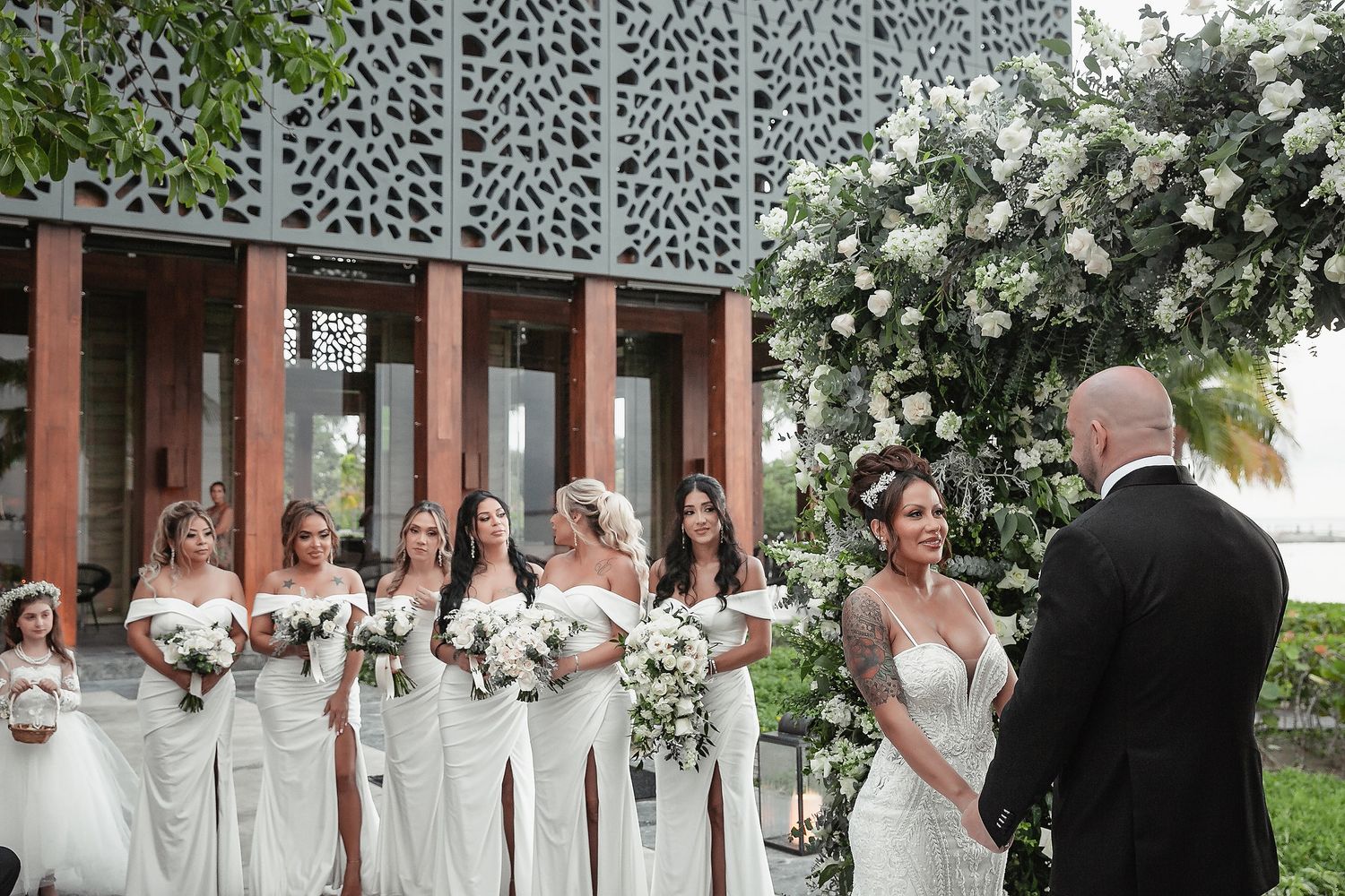 Wedding party in white dresses gathers under decorative lattice wall with white floral arrangements during outdoor ceremony.