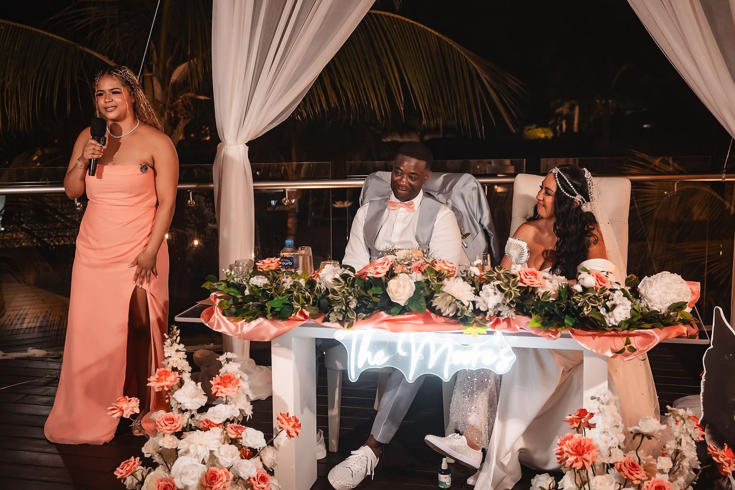 A wedding reception setup with coral floral decorations and white draping under string lights.