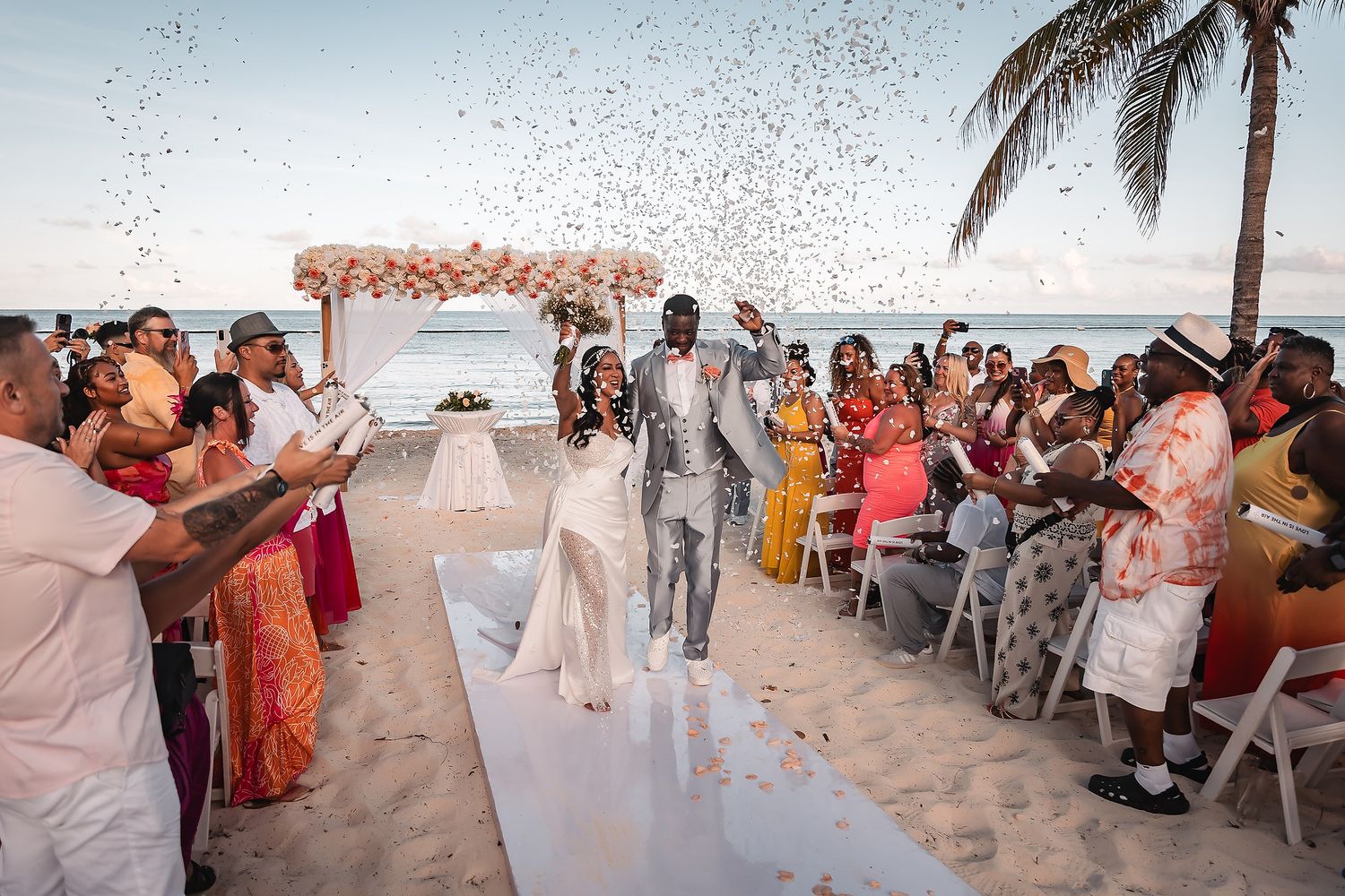 A romantic beach wedding ceremony under a floral arch with guests seated on white chairs by the ocean.