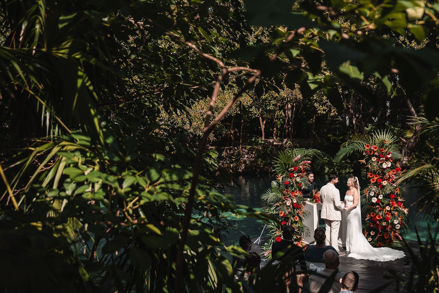 Wedding ceremony viewed through tropical foliage with red floral arch decorations.