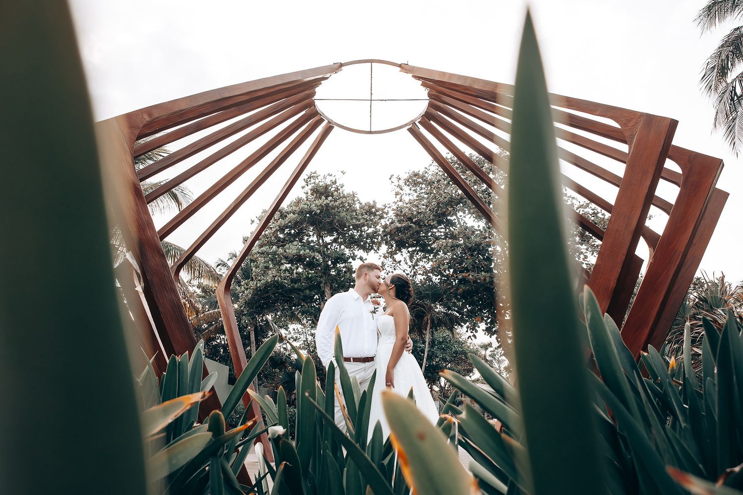A wedding photo captured through foliage under a geometric wooden pergola with palm trees.
