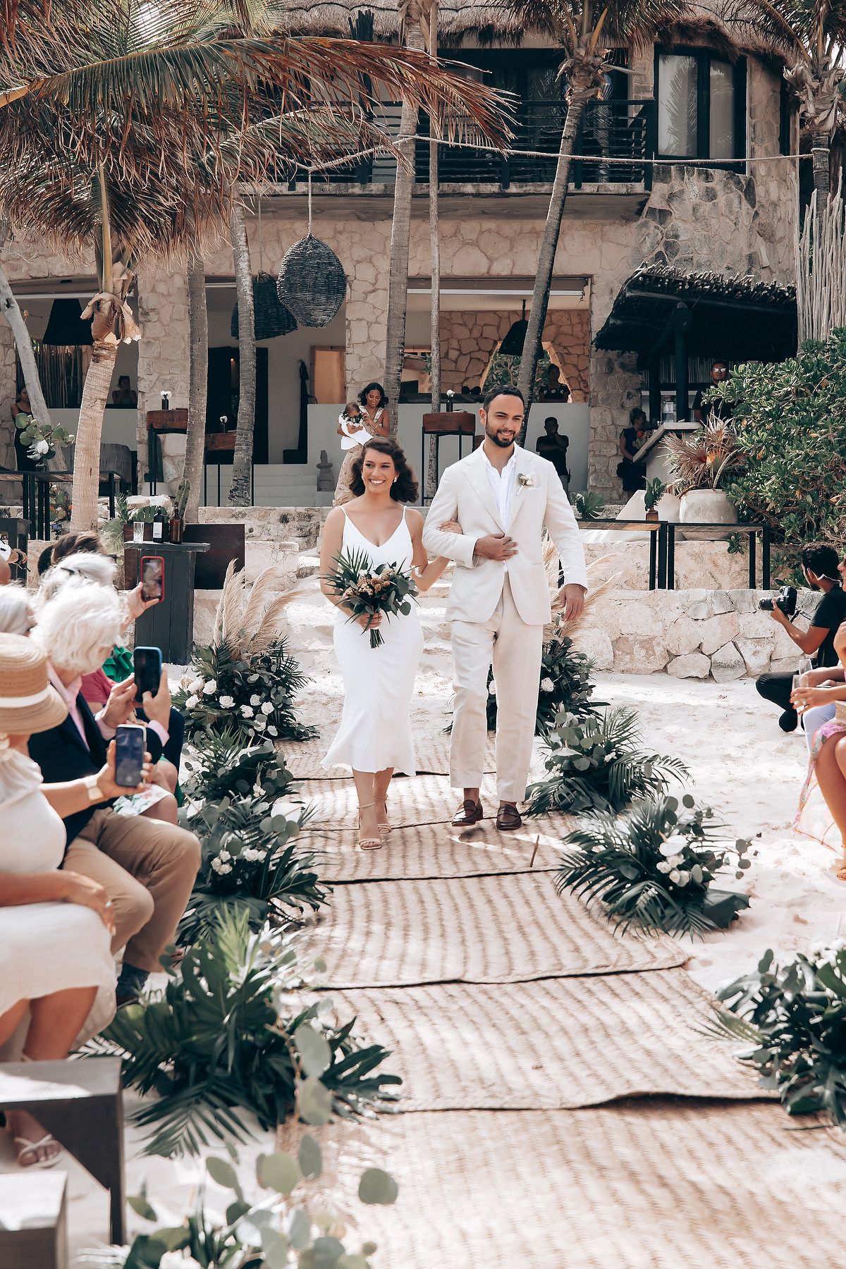 Wedding ceremony procession on stone pathway lined with tropical flowers and palm trees.