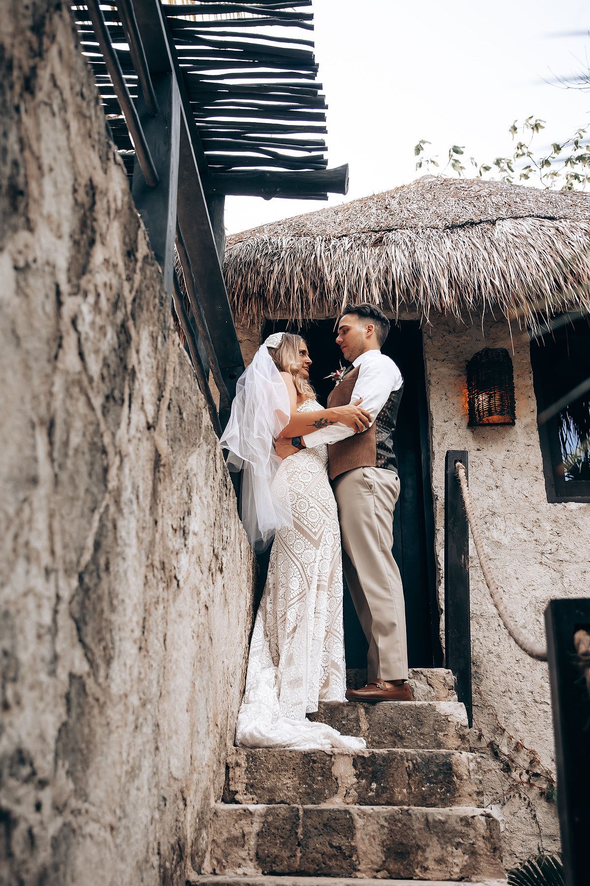 A romantic wedding moment captured against a rustic thatched-roof building with stone walls.