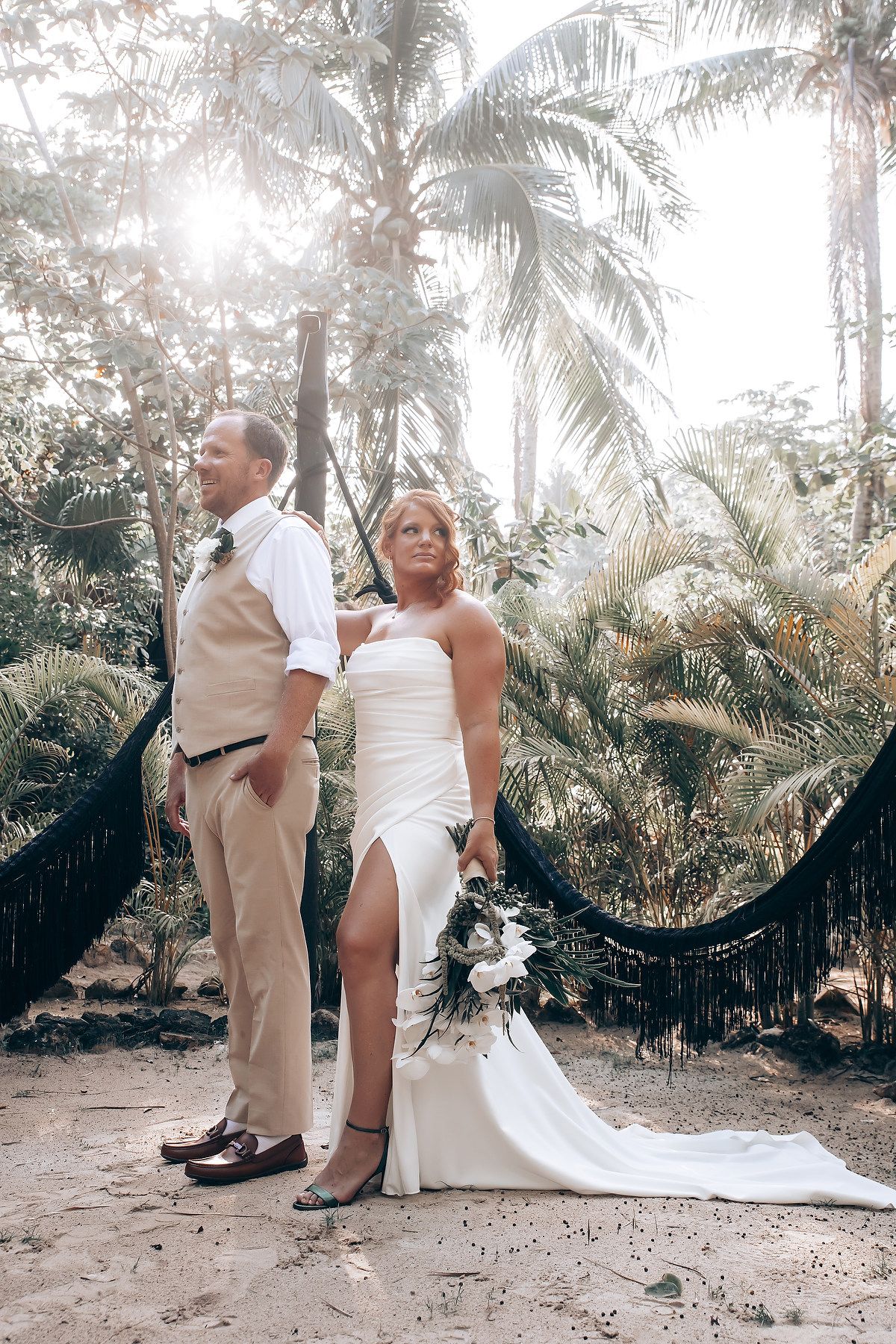 Newlyweds in white and beige attire pose elegantly against a backdrop of palm trees.