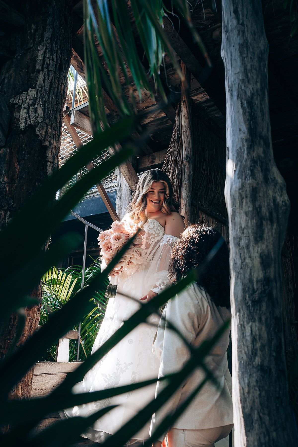 Intimate wedding photography captured in a shadowy wooden structure with palm fronds.