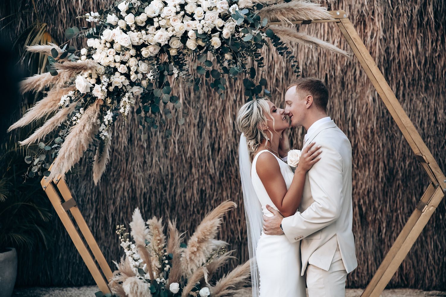 A couple sharing a romantic moment under a hexagonal arch decorated with dried flowers and pampas grass.