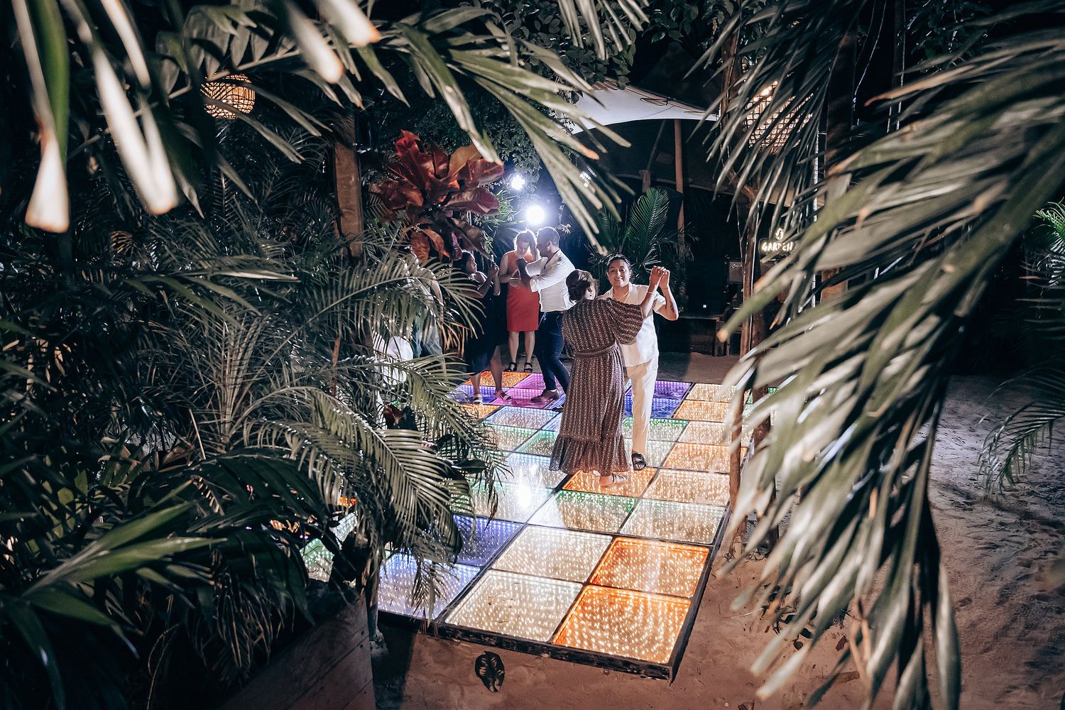 Partygoers dancing on an illuminated floor surrounded by tropical palm decorations.