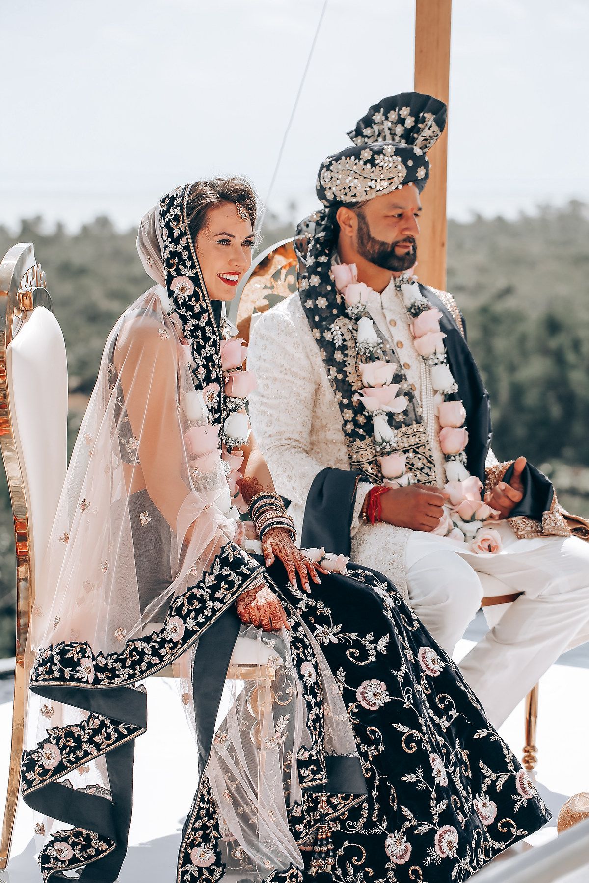 Couple in ornate black and white traditional wedding attire seated during outdoor ceremony.