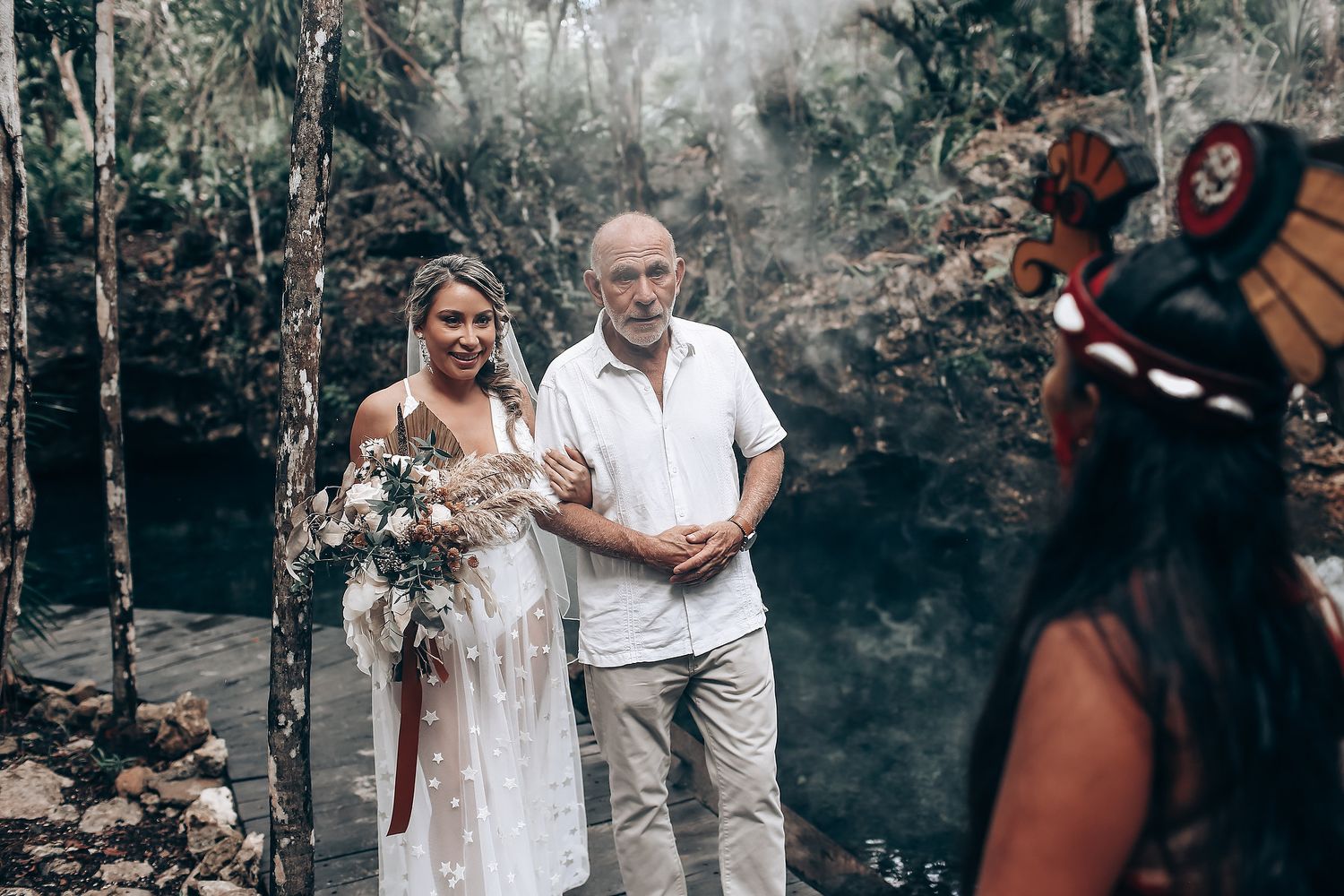 A couple in white attire embraces during an outdoor forest ceremony with stone walls in the background.