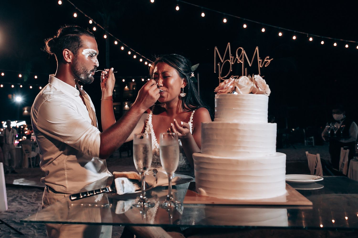 Wedding cake cutting ceremony under string lights at evening beach reception.