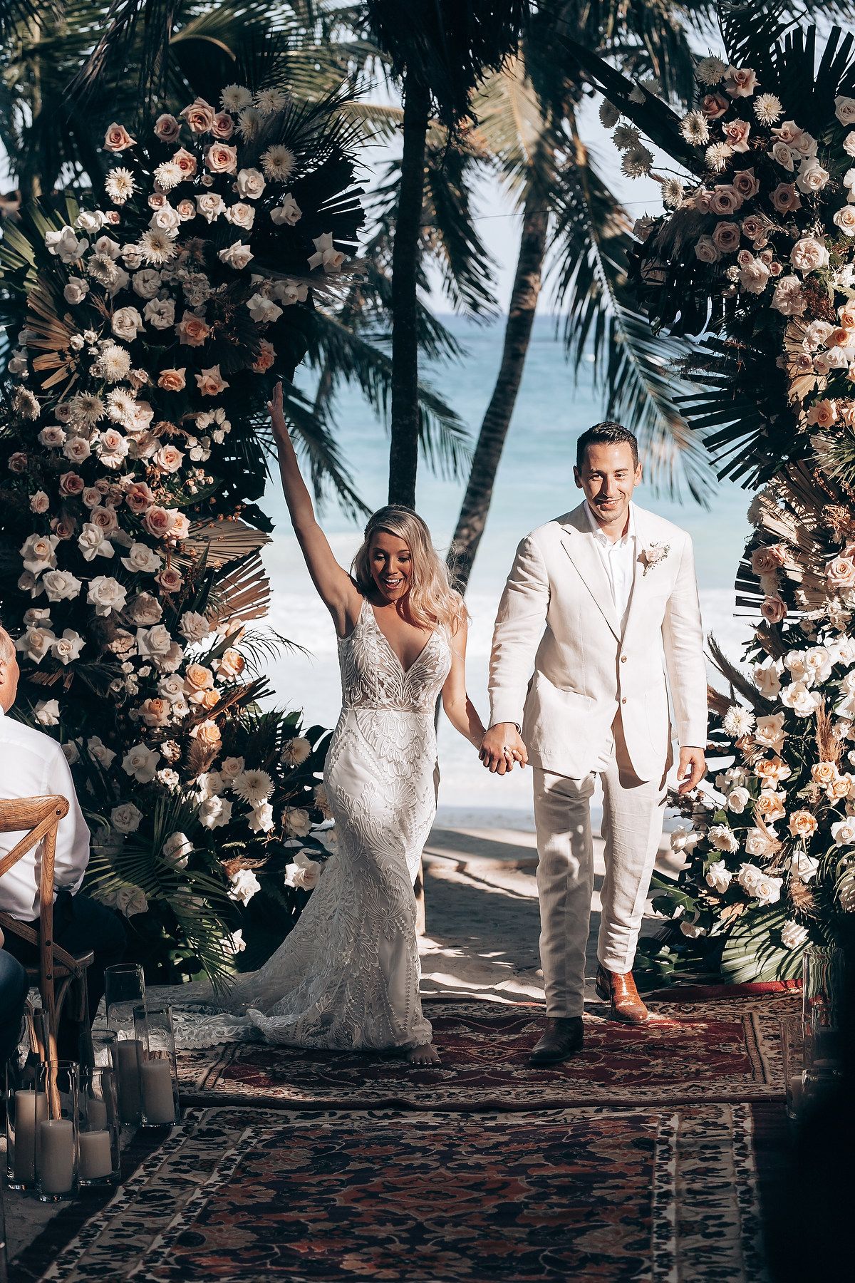 A wedding couple walks through a stunning floral arch with palm trees and ocean views.
