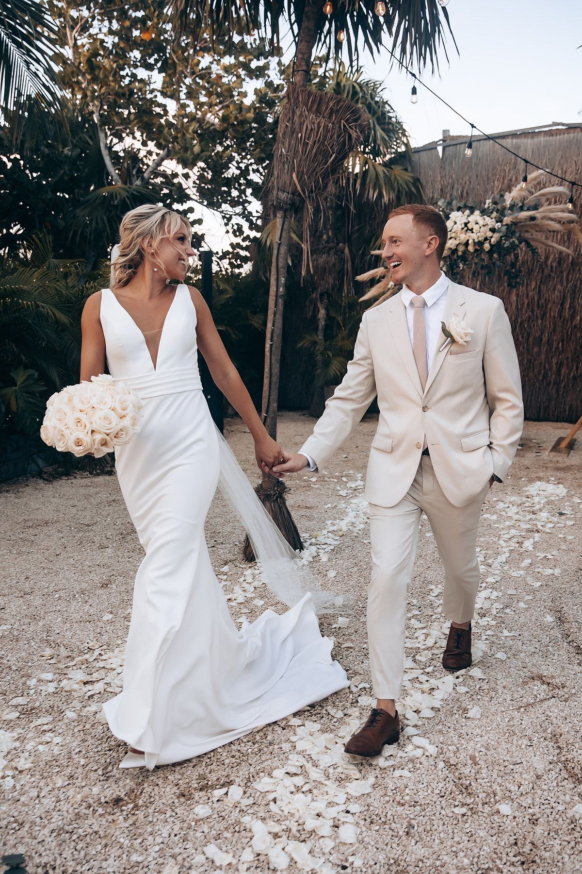 A couple in elegant white wedding attire dances playfully on a sandy beach path surrounded by palm trees.