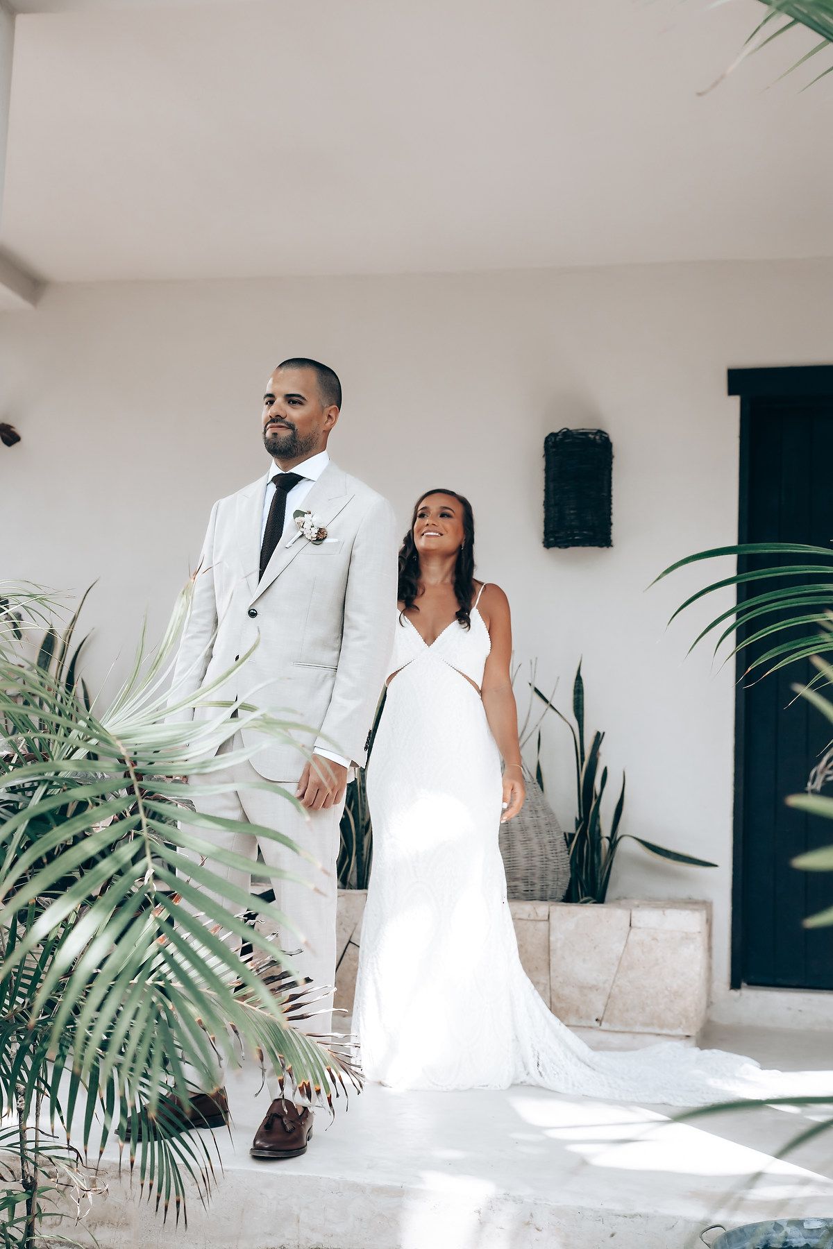 Couple in white wedding attire poses against a modern white wall with tropical palm leaves.