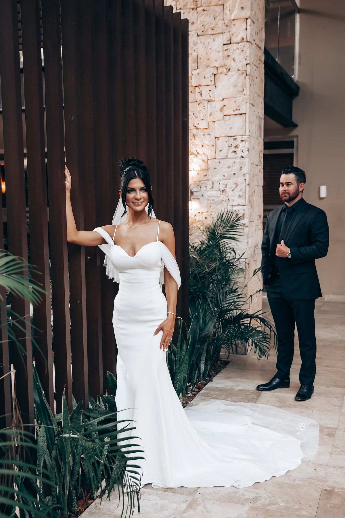 A bride in a white wedding gown reveals herself during a first look moment against a stone wall backdrop.