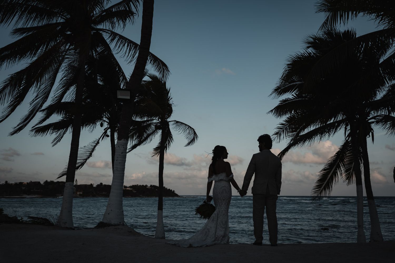 Silhouette of bride and groom holding hands under palm trees at dusk on tropical beach at sunset.