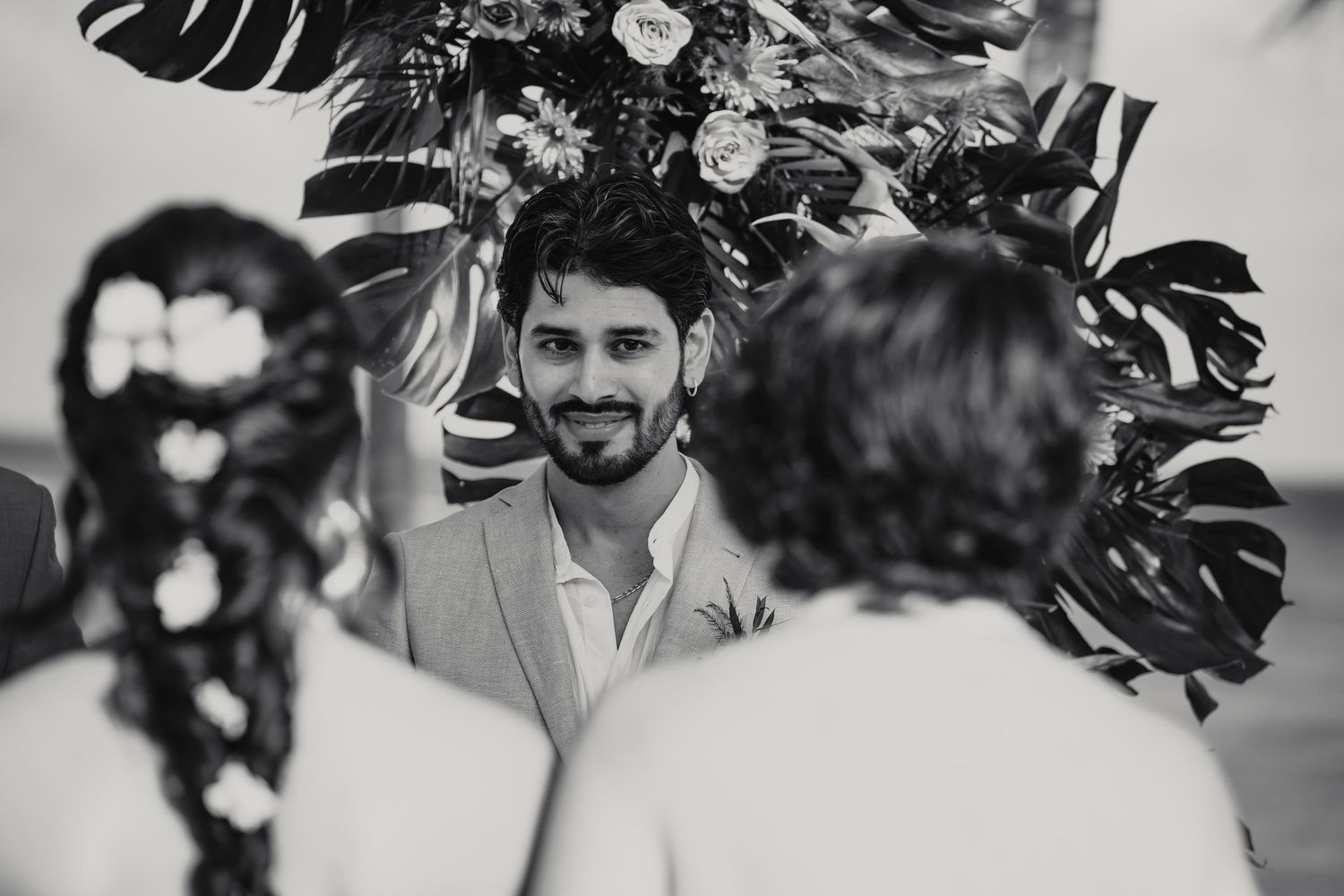 Groom smiling warmly at bride during outdoor beach wedding ceremony with tropical monstera leaf floral arch.