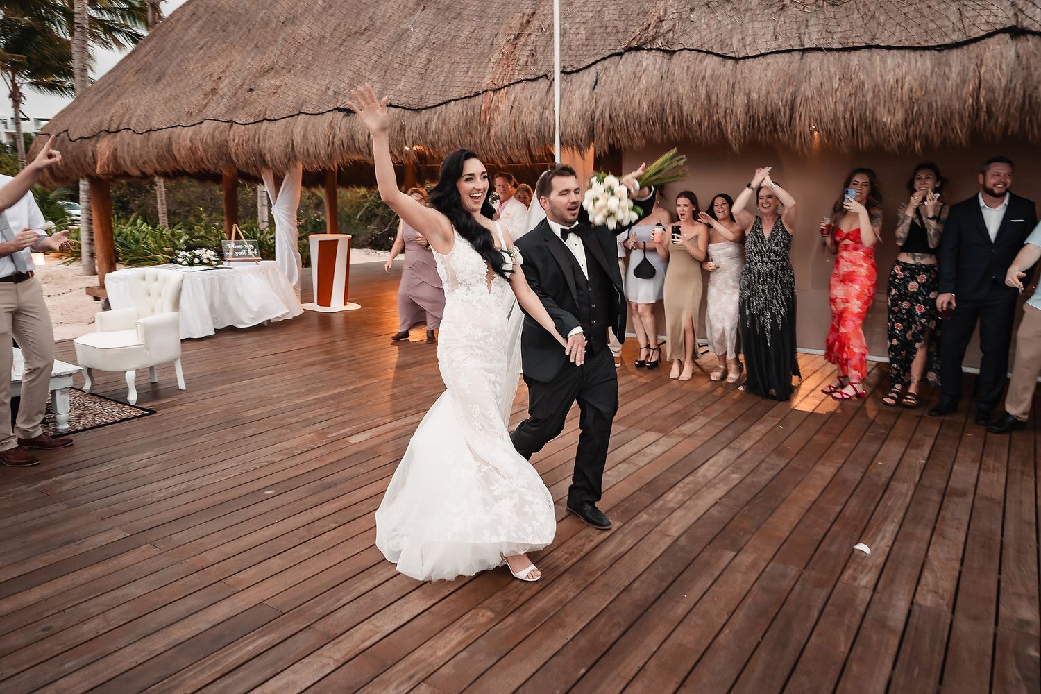 Bride and groom make grand entrance on tropical wooden deck at outdoor wedding reception with guests cheering.