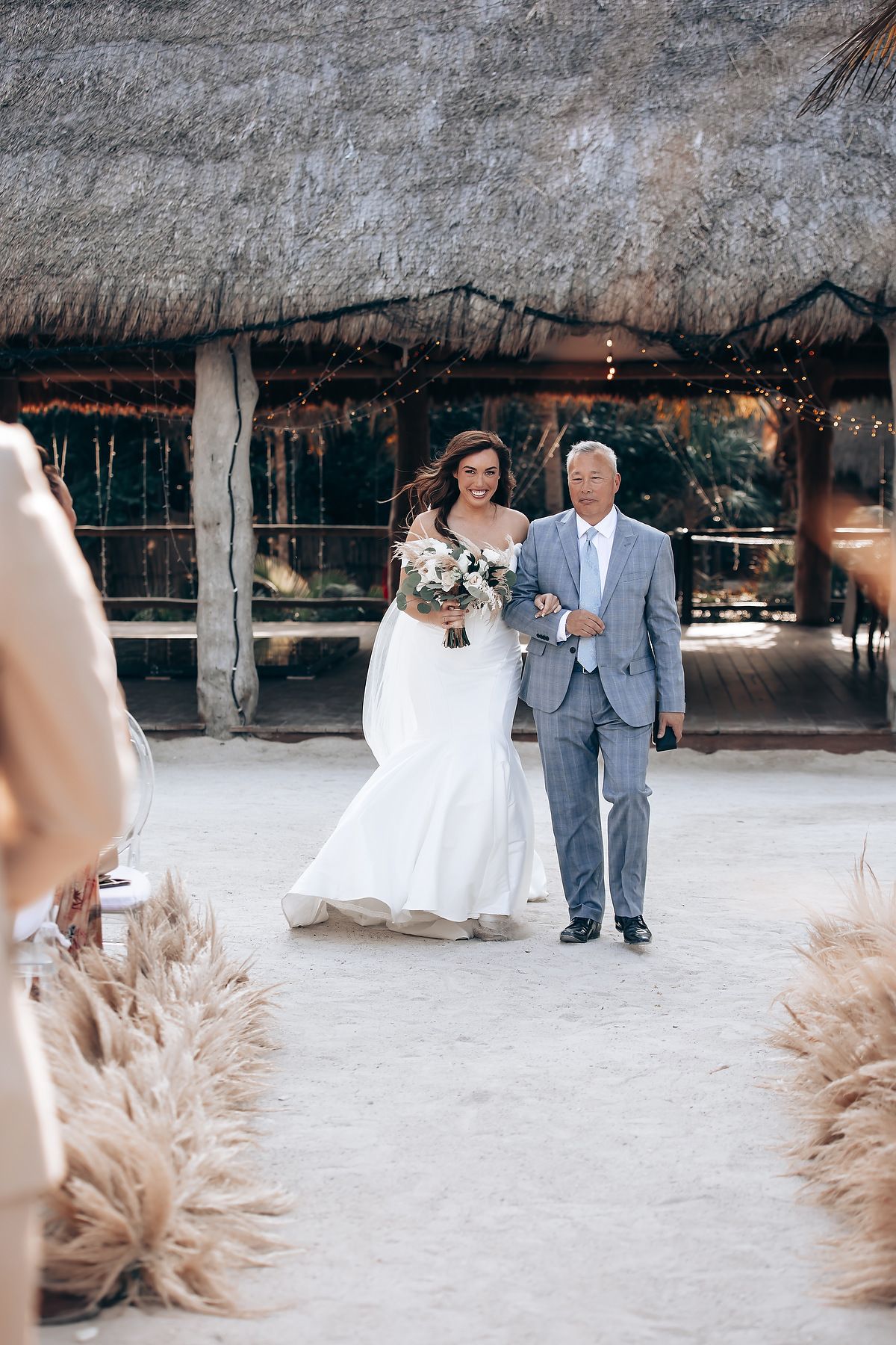 A panoramic view of a beach wedding ceremony setup with thatched roof and pampas grass decorations on sandy ground.