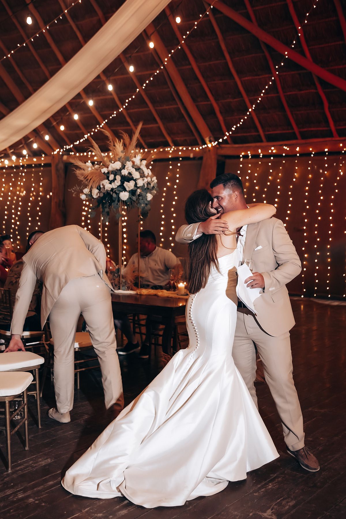 A couple in white wedding attire dances together under twinkling string lights in a rustic barn venue.