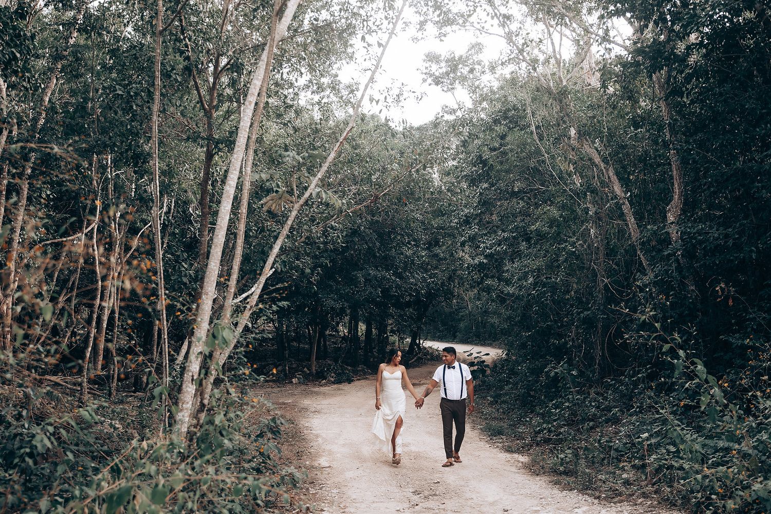 A couple walks hand in hand along a serene forest path surrounded by lush green trees.