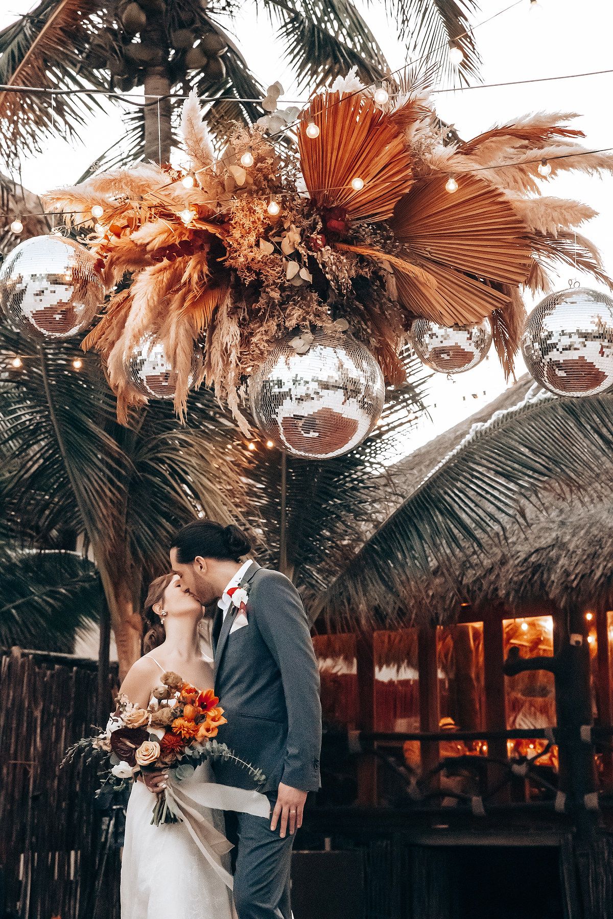 A romantic wedding couple shares intimate moments under palm fronds and hanging glass orbs at a tropical venue.