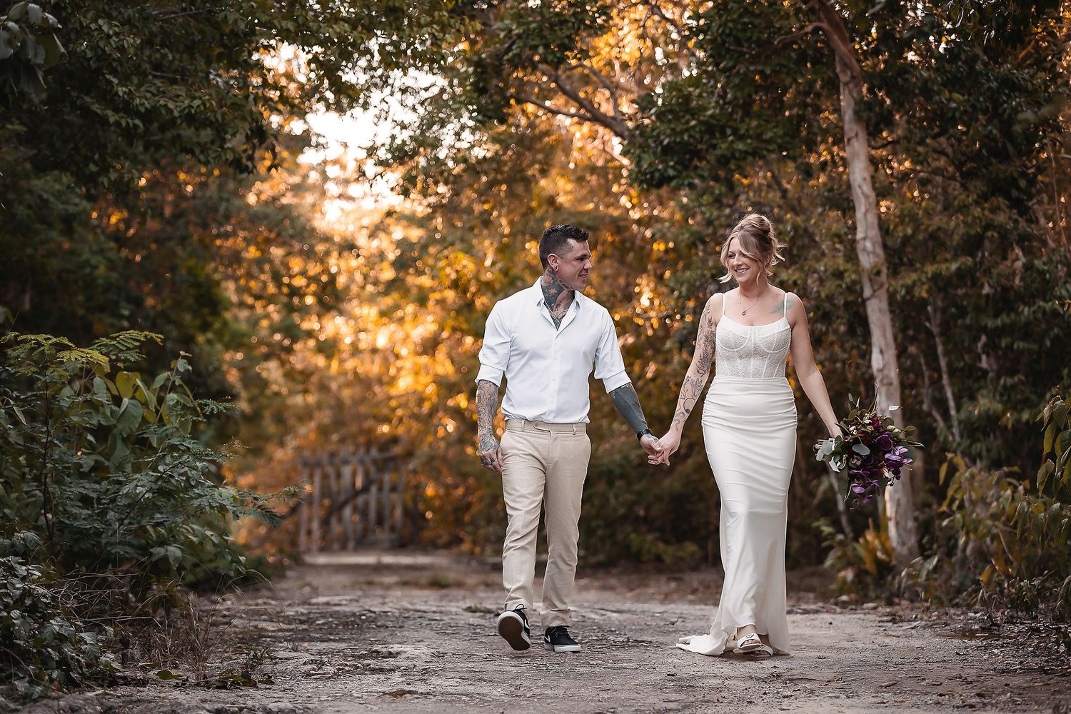 Smiling bride and groom stroll along a golden sunlit forest path holding hands.
