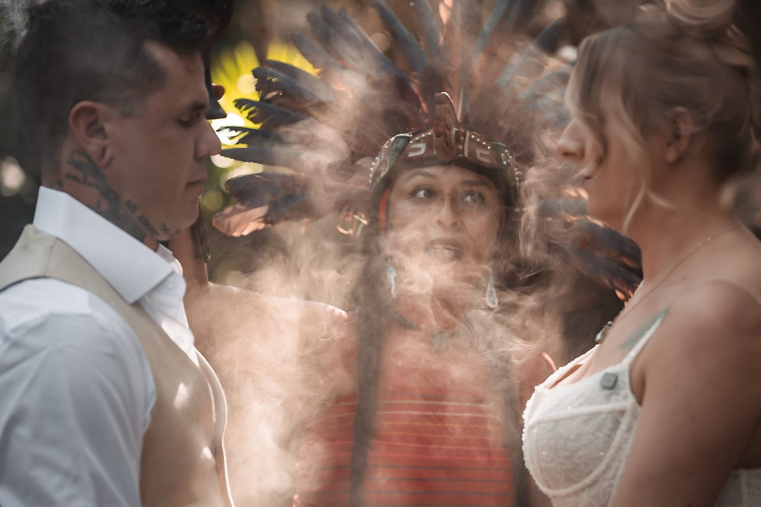 Native shaman in feathered headdress performing a smoke ceremony for a couple during an outdoor wedding ritual.