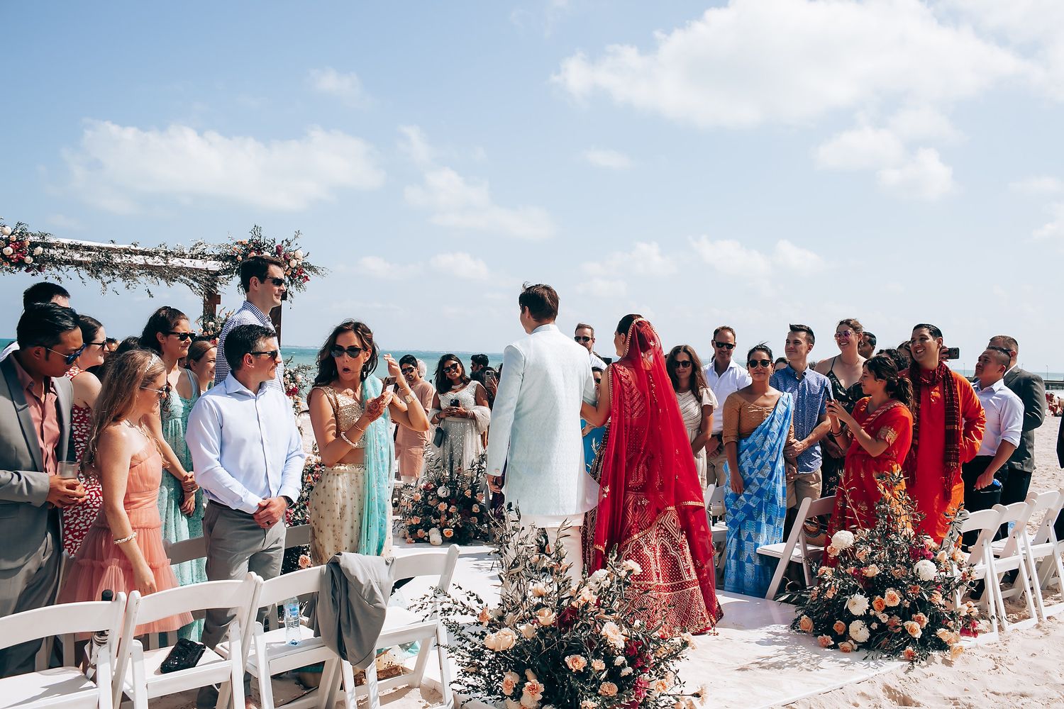 Wedding guests gather on a scenic beach for an outdoor ceremony with ocean views and floral decorations.