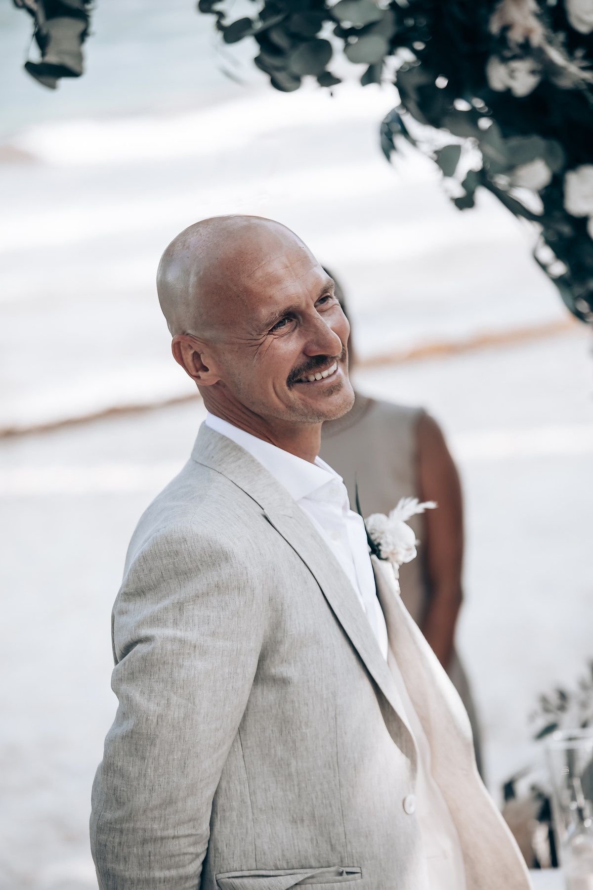 A joyful groom in a light beige suit and white boutonniere smiles during an outdoor ceremony.