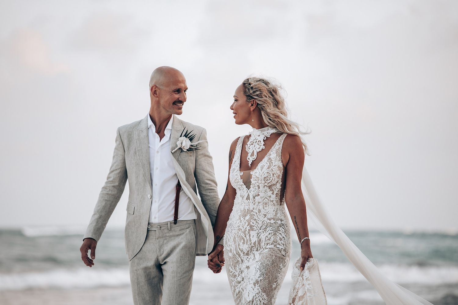 Couple walking along beach shoreline in elegant wedding attire with waves in background.