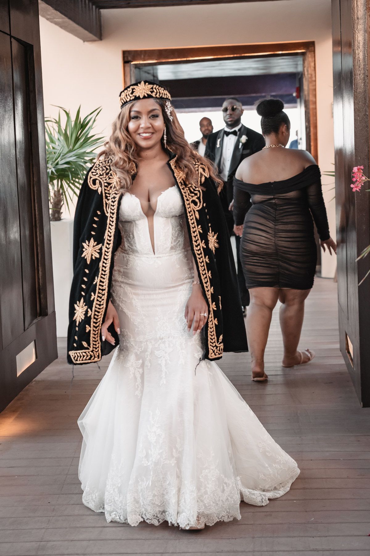 Bride in mermaid lace gown with ornate black and gold cape headpiece at wedding entrance.