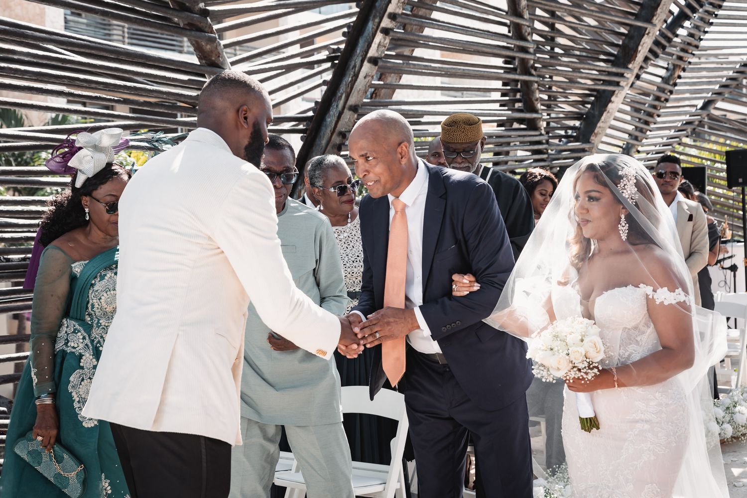 Groom in white suit greeting guest while bride in lace gown walks down aisle.