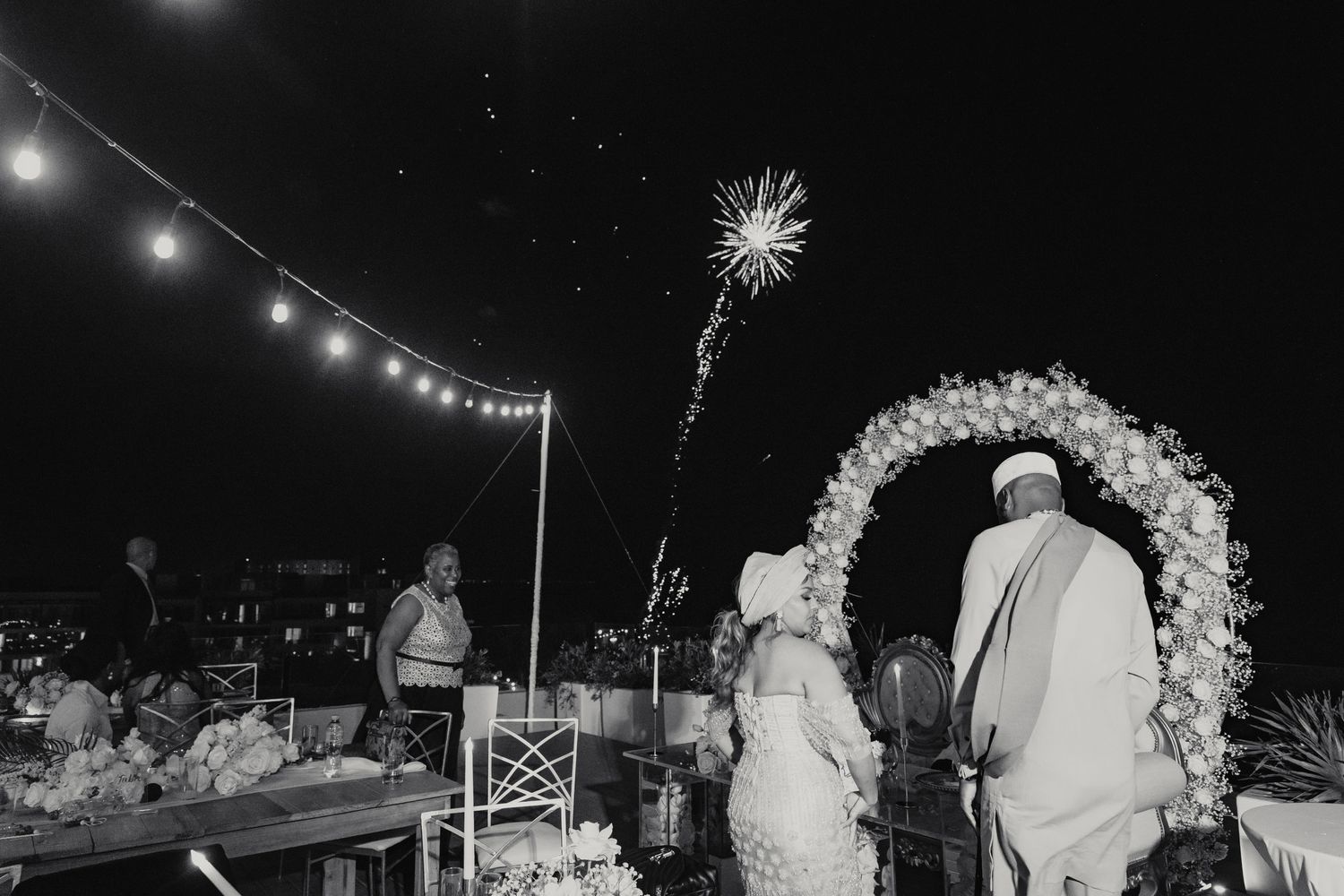 Bride and groom stand beneath a floral arch as fireworks sparkle at their outdoor night wedding.