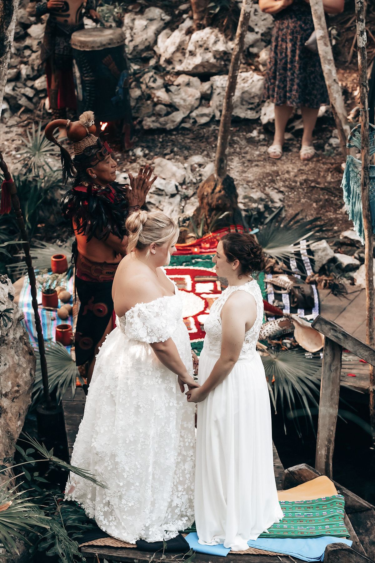 Two brides in white wedding dresses hold hands in a lush, tropical setting.