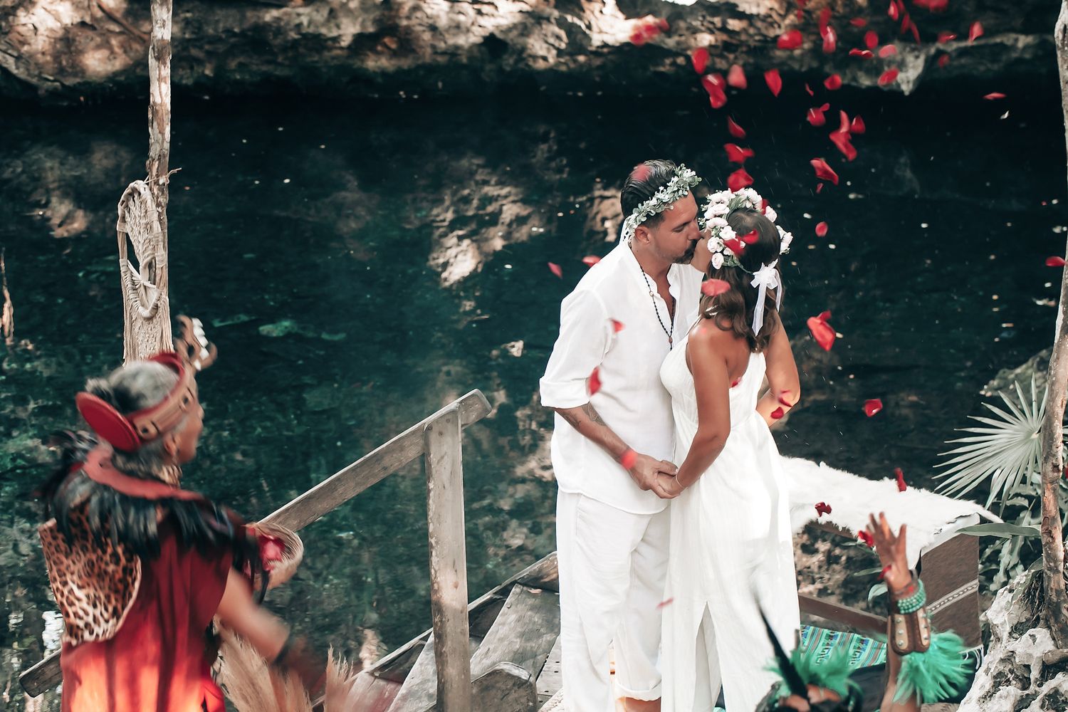 A couple in white clothing stands on a wooden bridge surrounded by flowers and greenery.