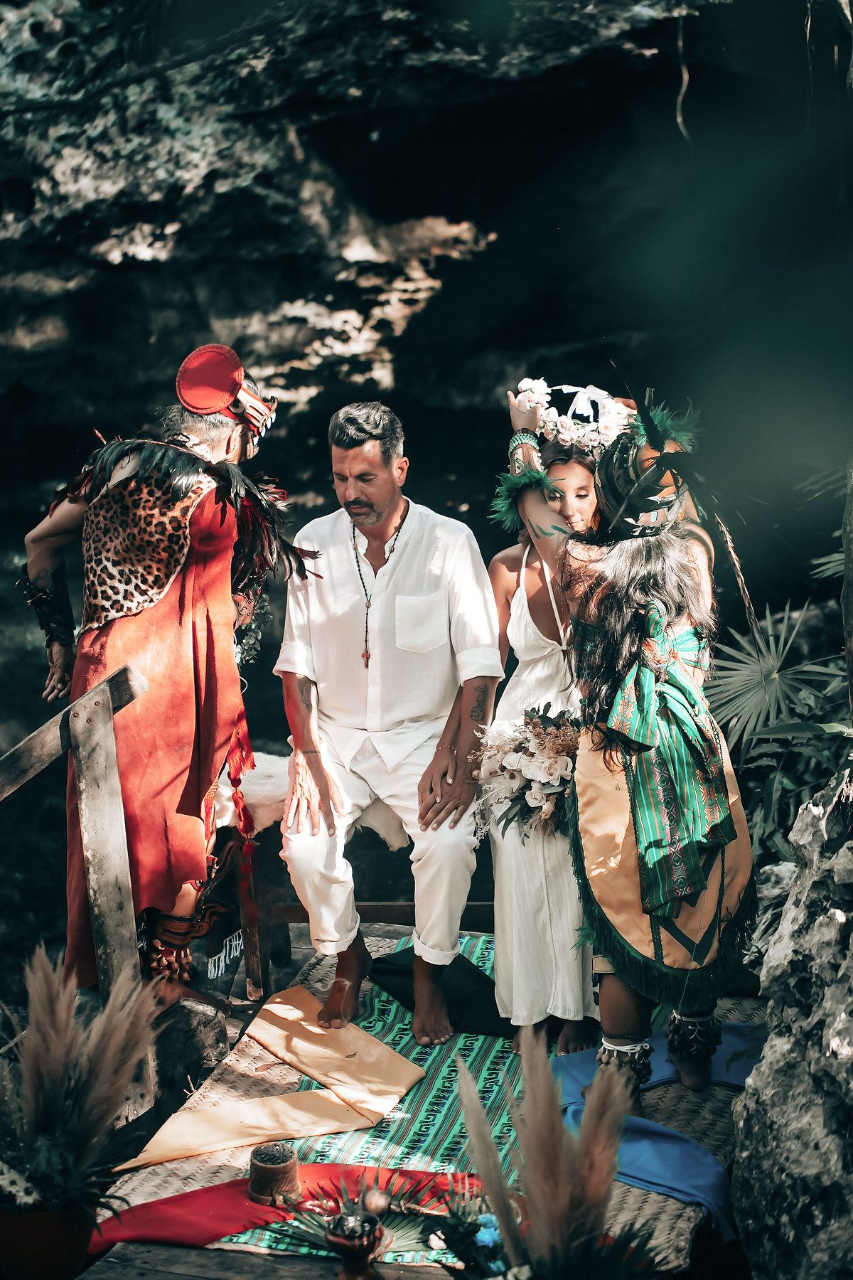 The image depicts a group of people wearing colorful, ornate costumes and standing on a wooden platform in a lush, tropical setting.