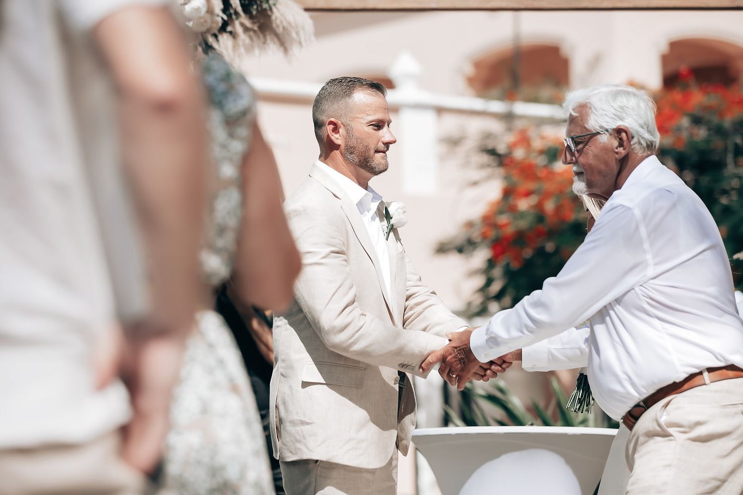 Two men shake hands at a wedding ceremony, one in a suit and the other in a white shirt.