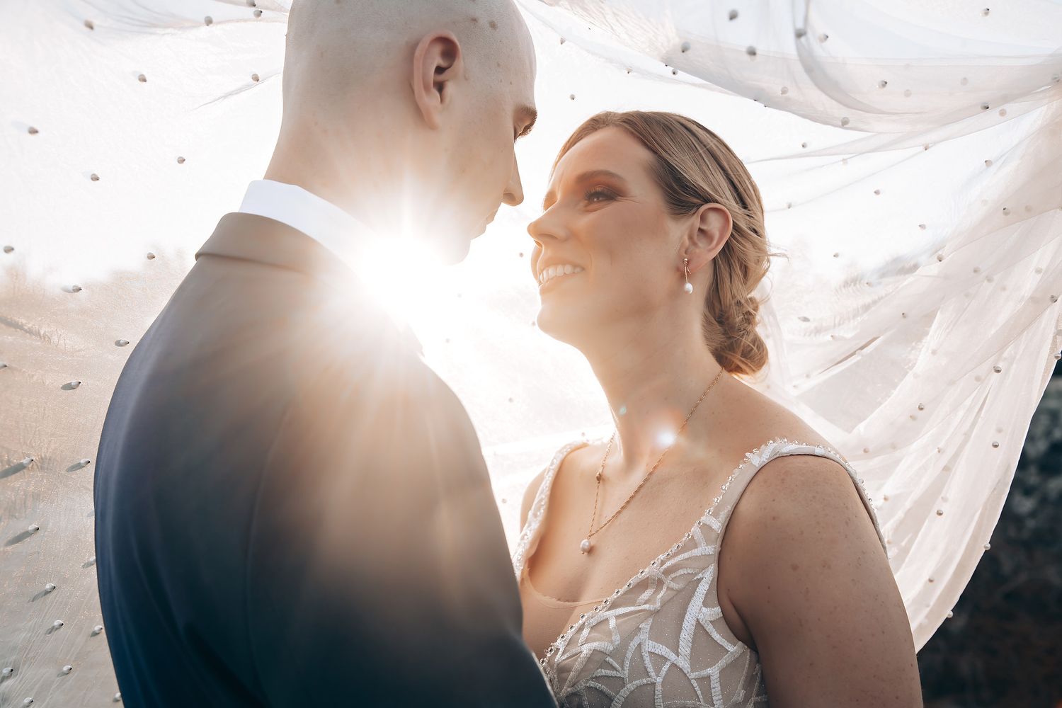 A bride and groom embrace in a romantic, backlit setting.