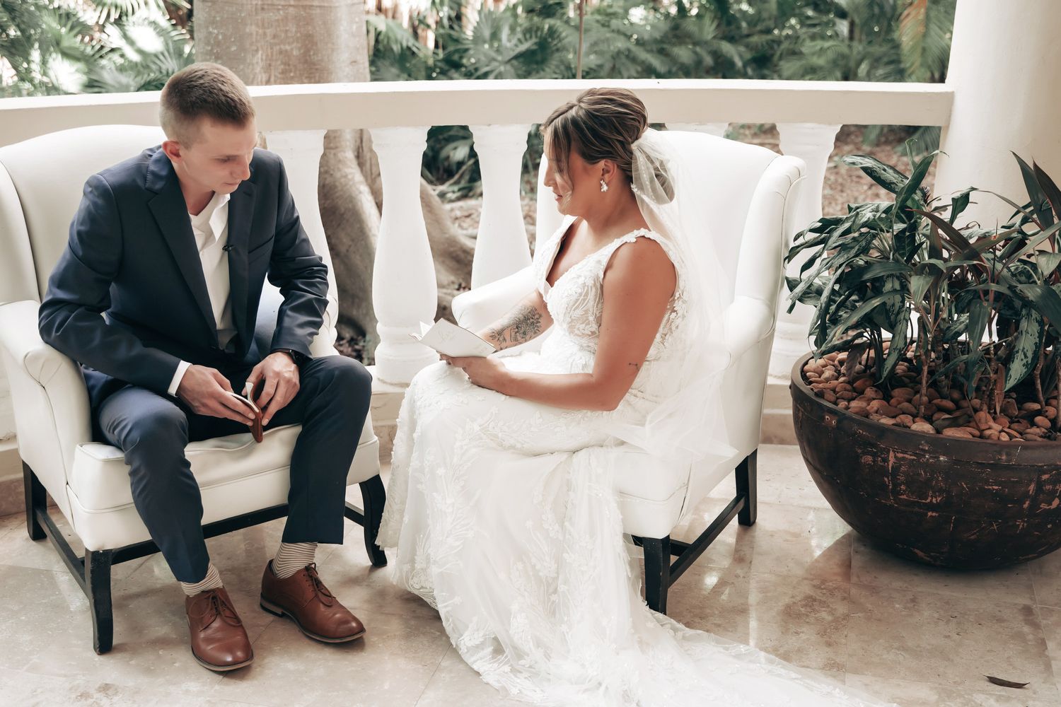 Bride and groom share a quiet moment seated in white chairs on an outdoor terrace before their ceremony.
