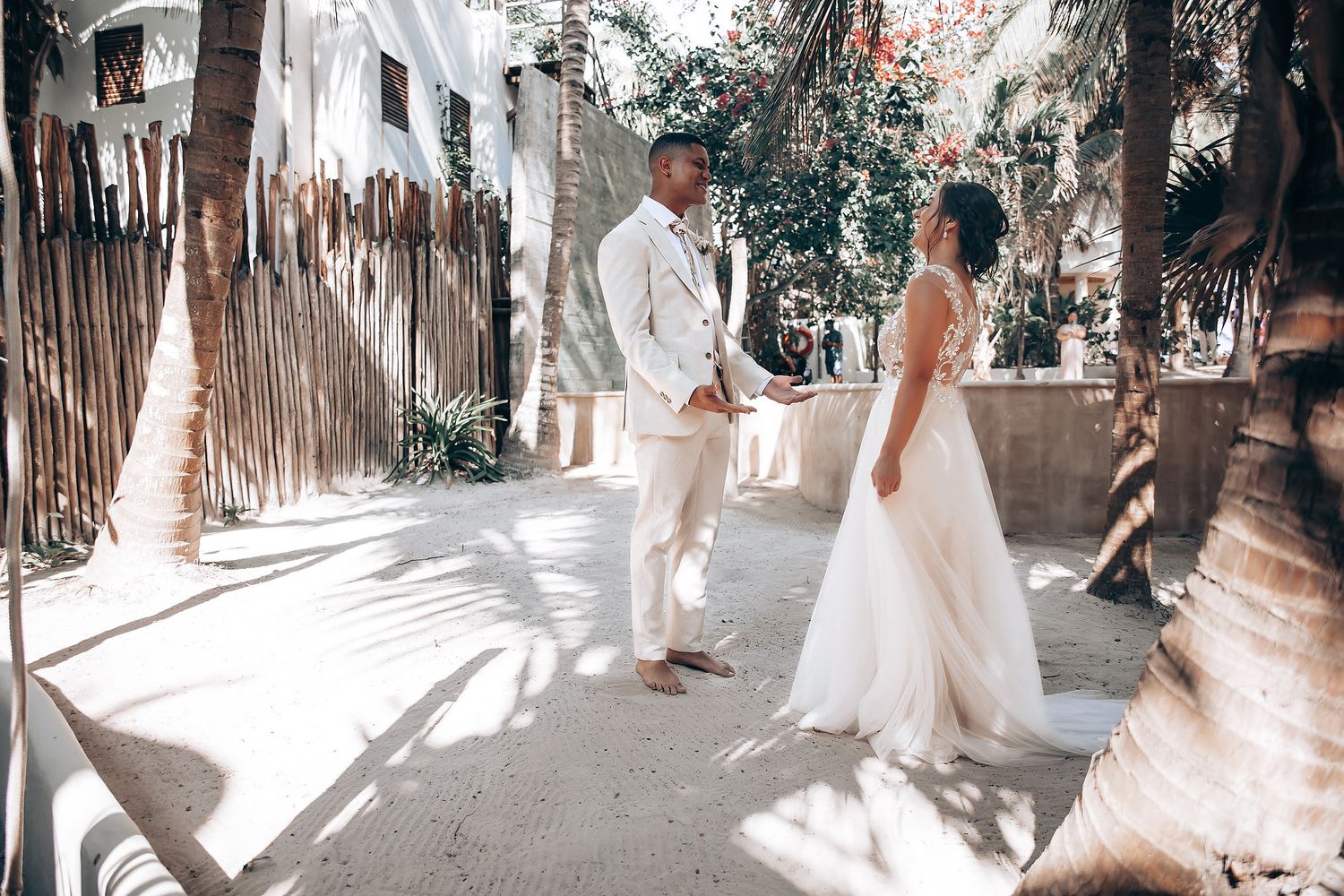 Wedding guests walk down a sunlit outdoor pathway lined with trees and bamboo fencing.