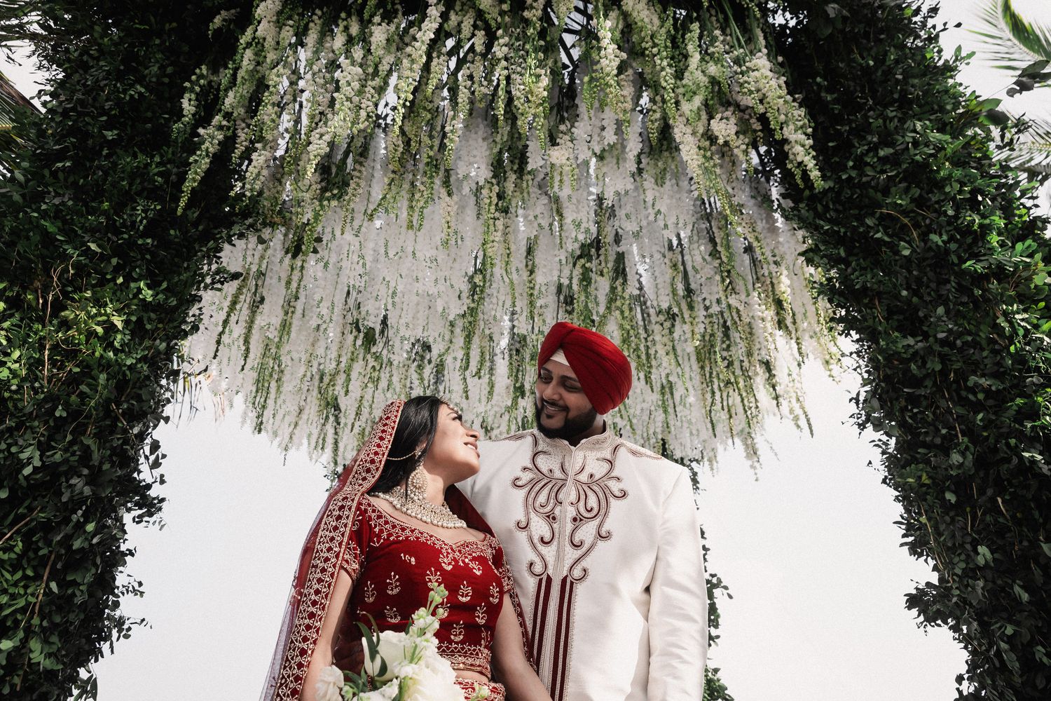Indian bride and groom in red and white traditional wedding attire gazing at each other under lush greenery floral arch.
