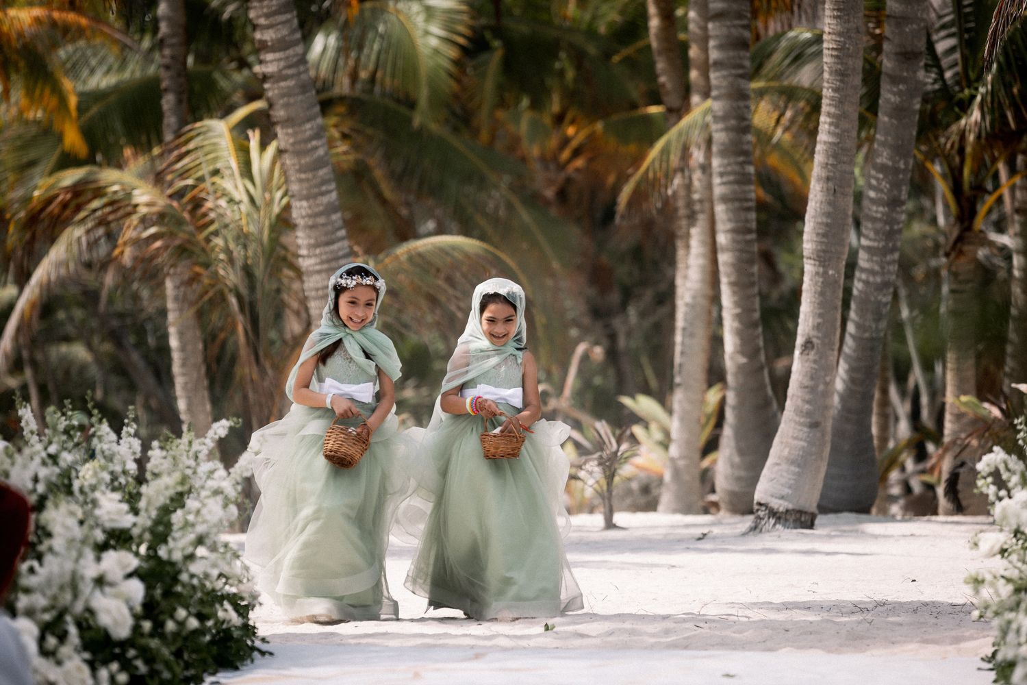 Two flower girls in mint green lehengas walking down tropical beach wedding aisle with palms.