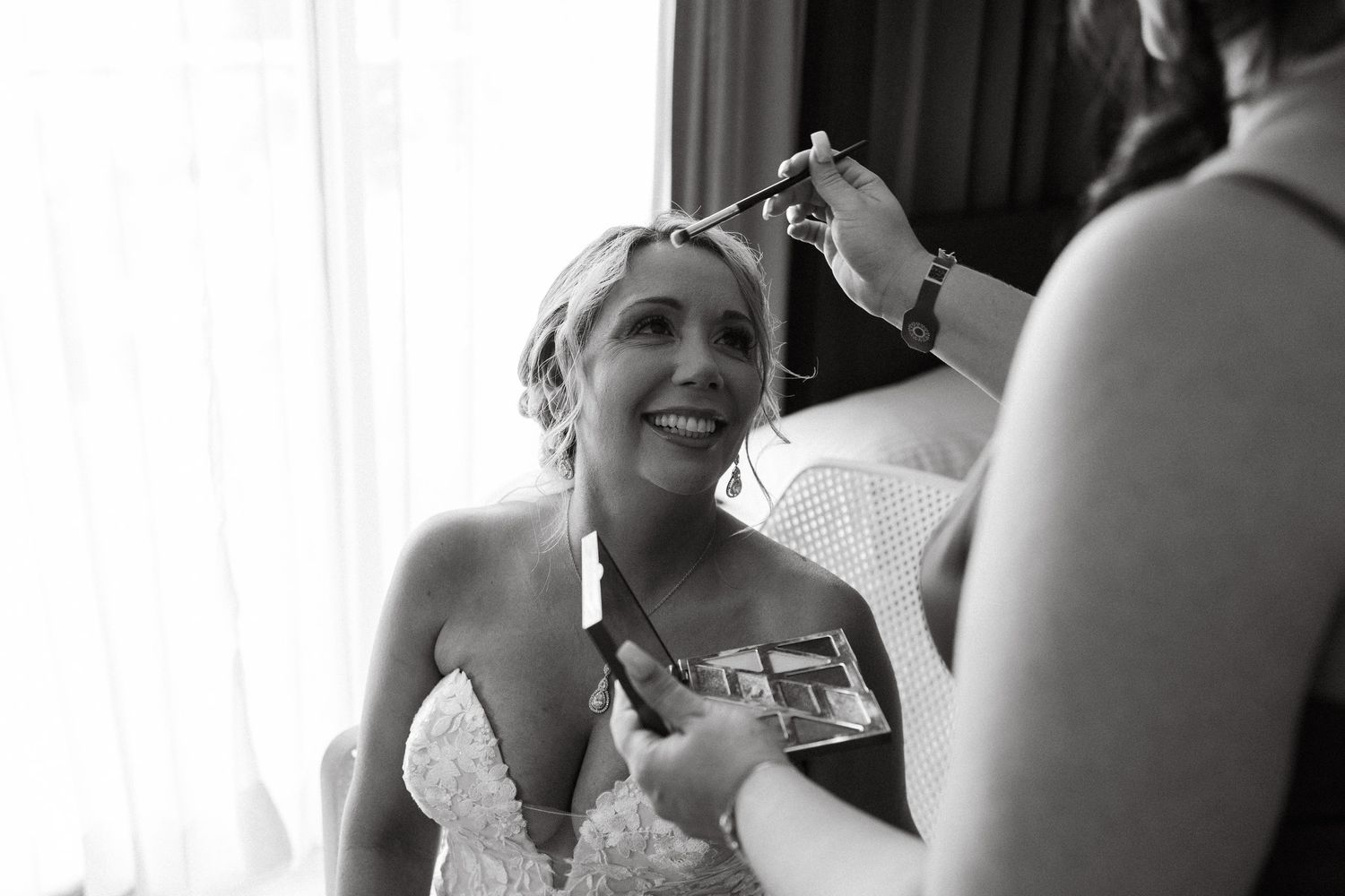 Bride smiling as bridesmaid styles her hair in black and white wedding preparation photo.