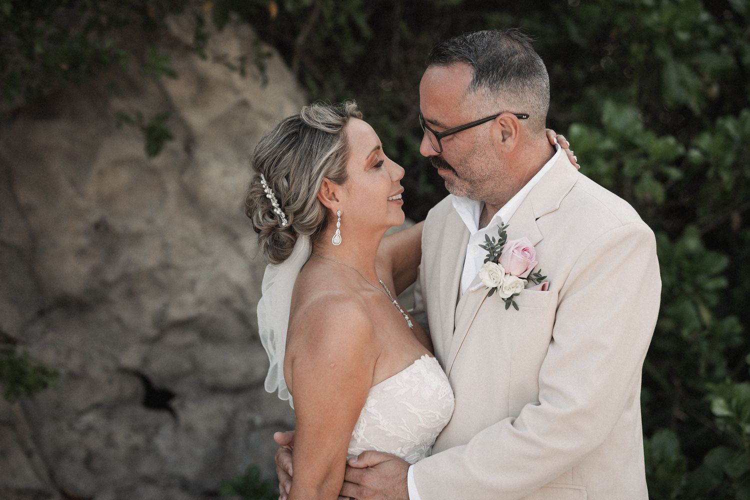 Bride in strapless lace gown and groom in cream suit share an intimate gaze during outdoor wedding ceremony.