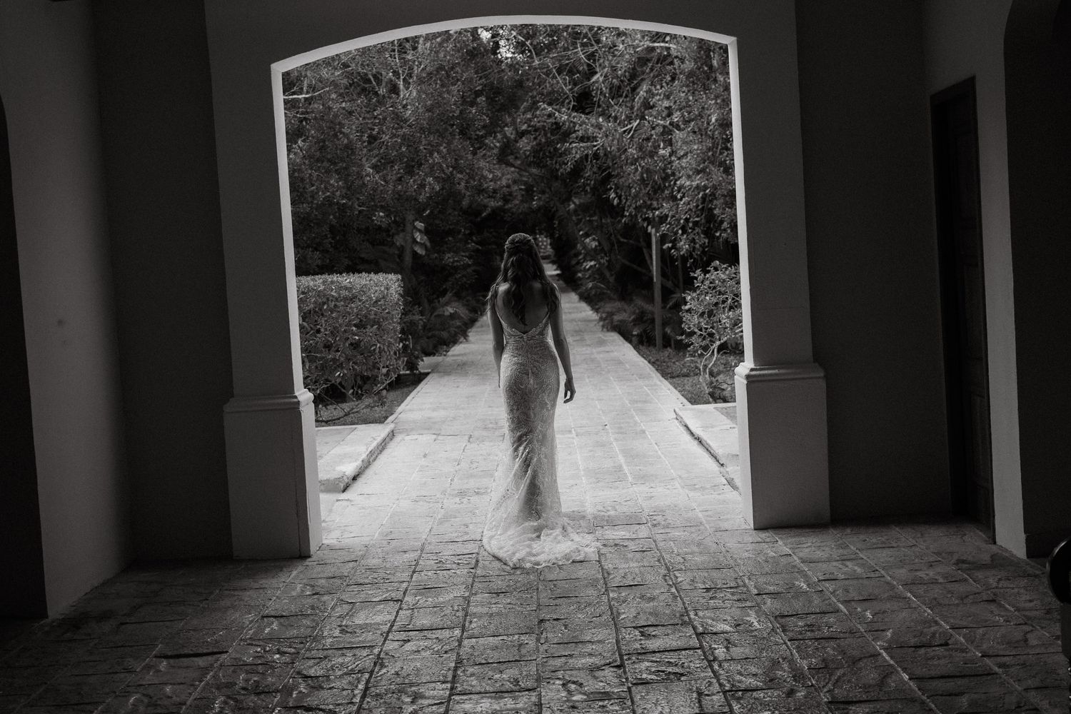 Black and white photo of a bride in a lace gown walking through an elegant stone archway.