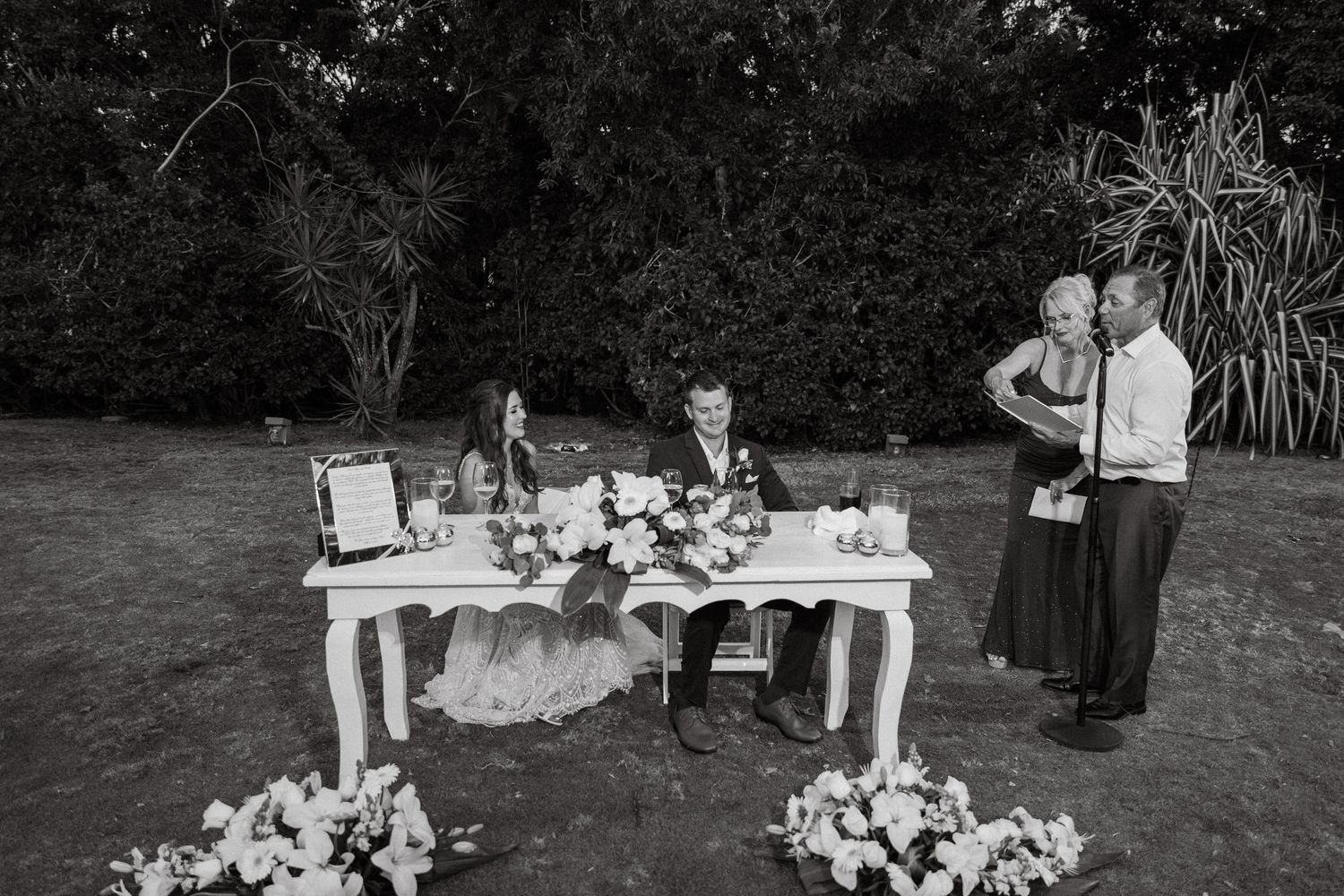 Bride and groom sign wedding documents at floral-decorated table while guests embrace nearby in black and white photo.